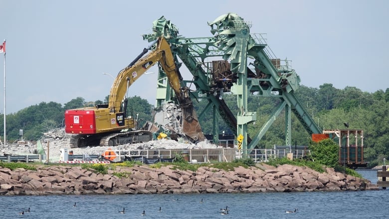 A large piece of heavy machinery is parked in front of a green, metal bridge that's torn apart. There's a large pile of concrete and twisted metal in front of it.