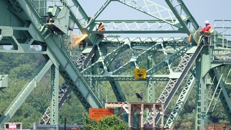 Sparks fly from the torches of two workers who are shown high up on the green metal structure of a bridge.