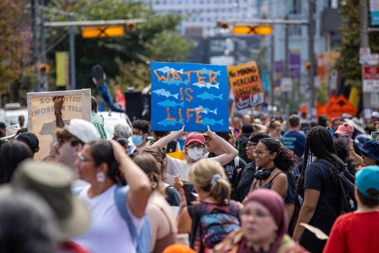 A person is seen in the centre of a large crowd outside, holding a sign that says "water is life."