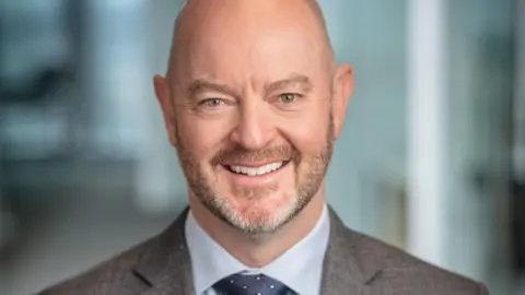 NexGen A professional headshot of Leigh Curyer smiling, wearing a grey suit jacket, a light blue button-up shirt and a speckled dark blue tie.