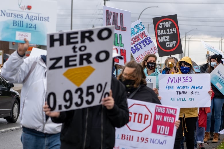 Protestors hold signs saying "Nurses are value greater than 0.9%" "Cease Invoice 124 wage lower" and "Hero to zero 0.935%"