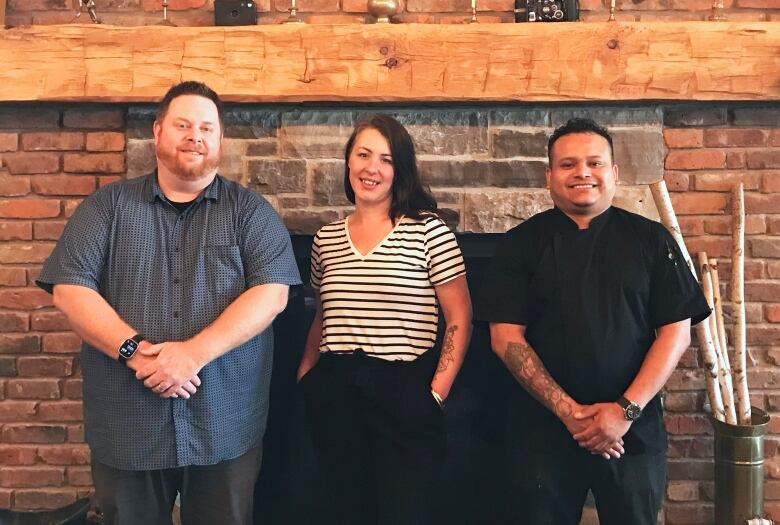 Three people stand in front of a fireplace.