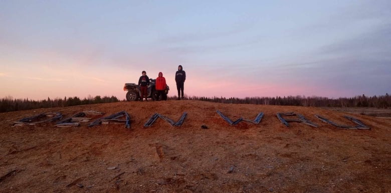 Three people are seen standing on a hill. A message is spelled out with pieces of wood that says "Dean WRU (where are you)."