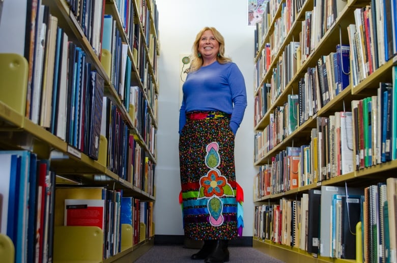 A person with blonde hair wearing a blue shirt and multi-coloured ribbon skirt stands in an aisle between bookshelves.