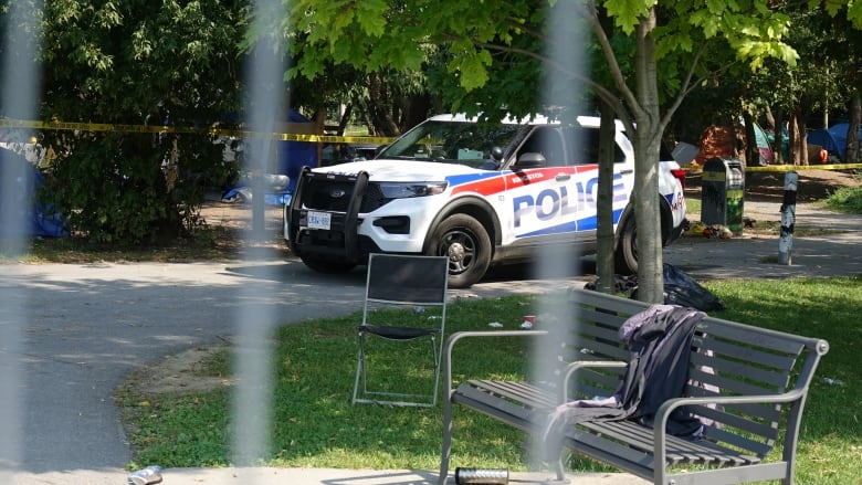 A police cruiser can be seen parked on a pathway, surrounded by tents, caution tape and an abandoned chair.