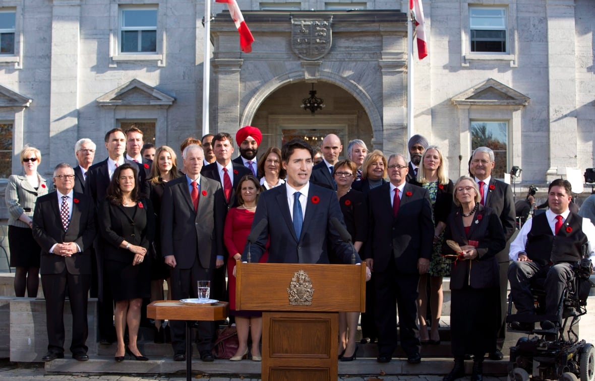 A politician speaks at a podium with roughly 30 people in suits and dressed behind him.