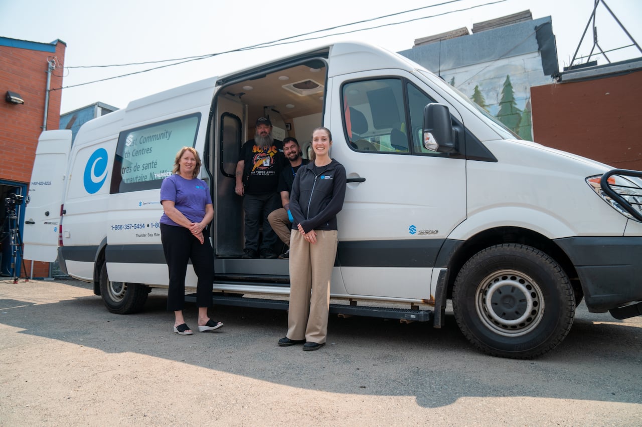 Two people are seen standing outside of a van with an open door, where two other people are seen inside.