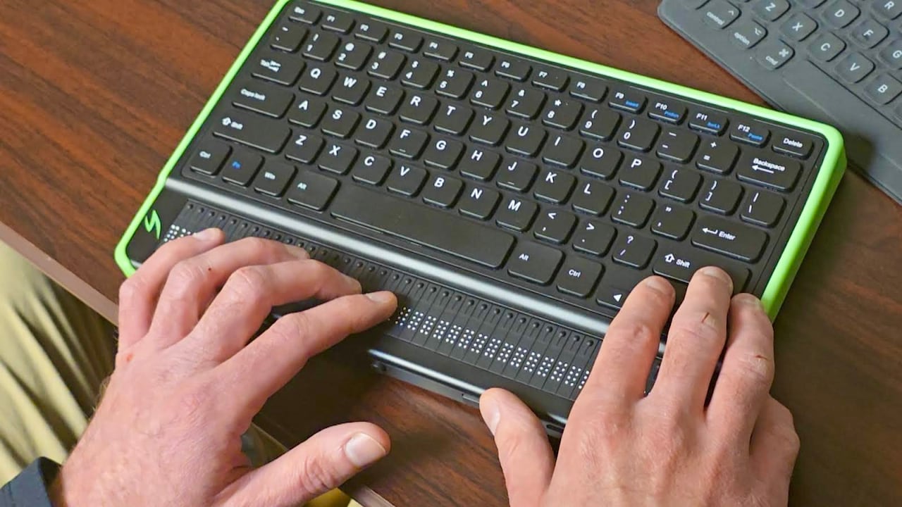 Two hands touching a braille keyboard sitting on a wooden desk. 