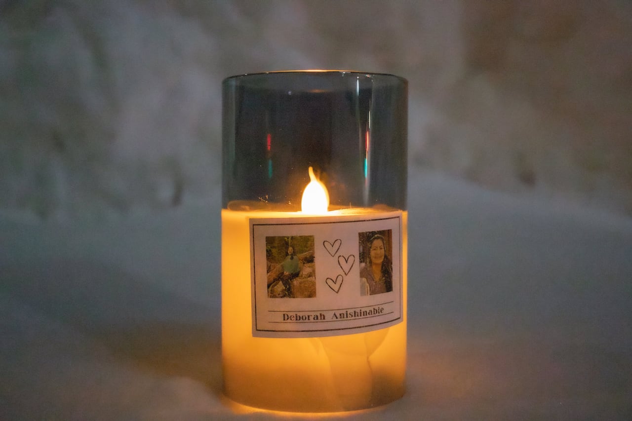 A candle is placed on the snowy ground. It has two photos of a woman on the front and says "Deborah Anishinabie."