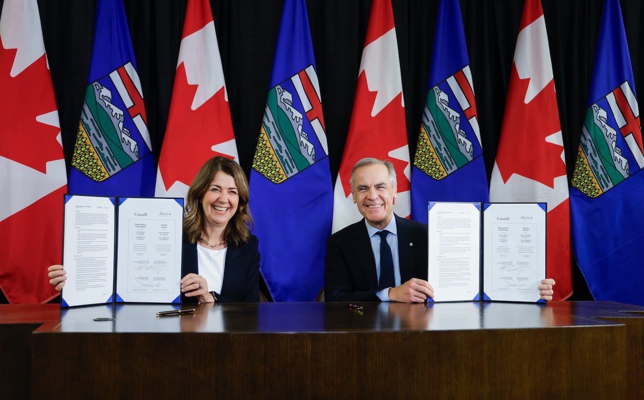 Mark Carney and Danielle Smith sit at a table, holding signed documents and smiling