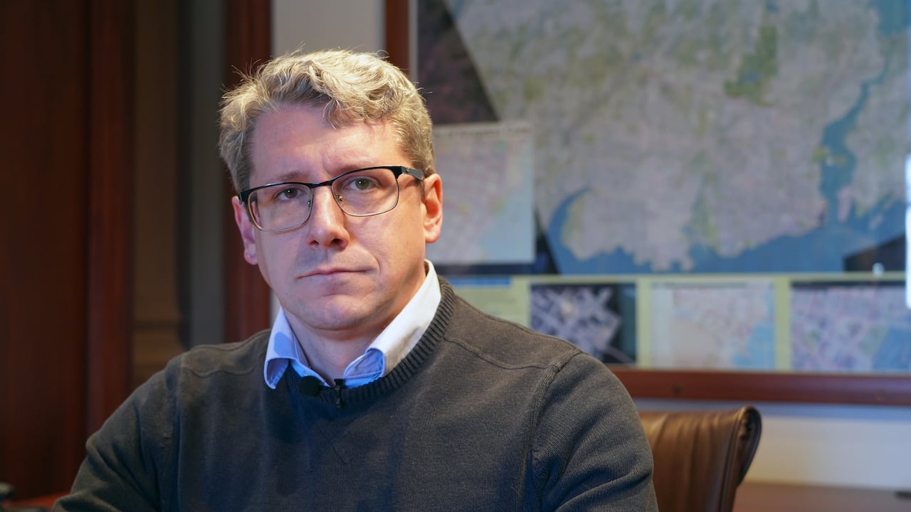 A man with grey hair and glasses sits in an office with a map of the City of Kingston on the wall behind him.