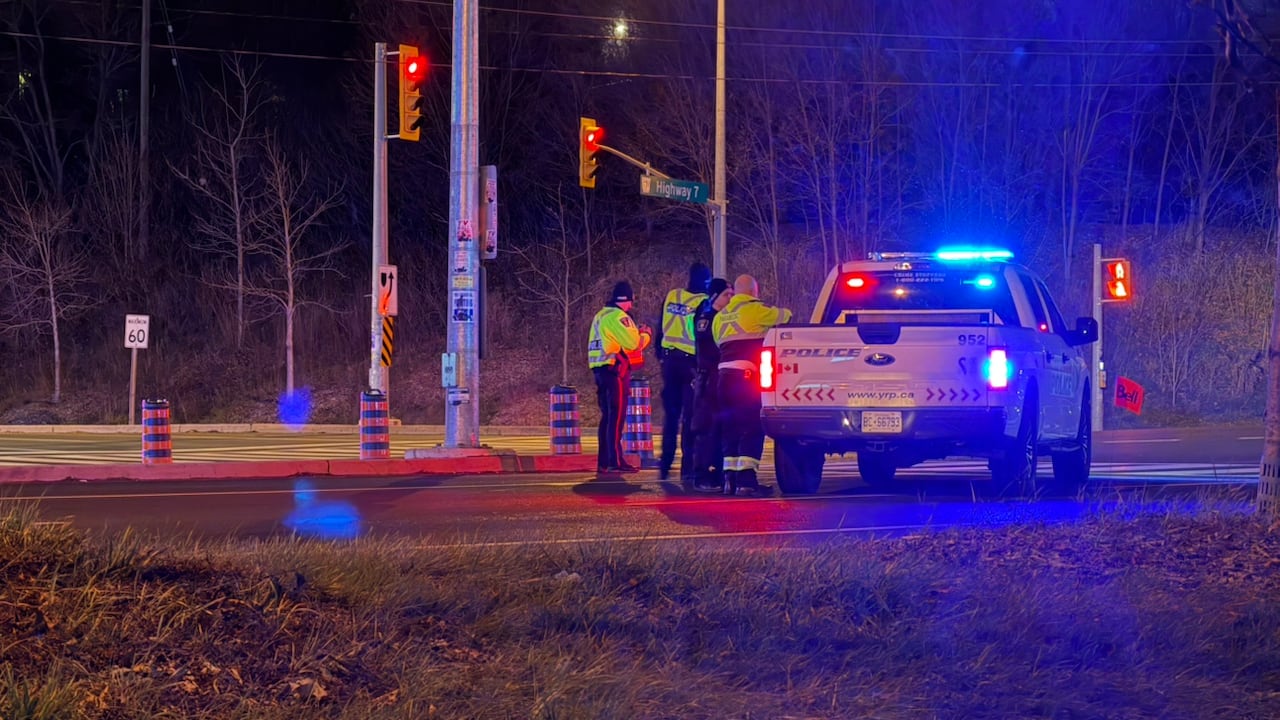 A police pickup truck is stopped at an intersection after dark. Four police officers are gathered, standing beside it. Three are wearing fluorescent safety vests.