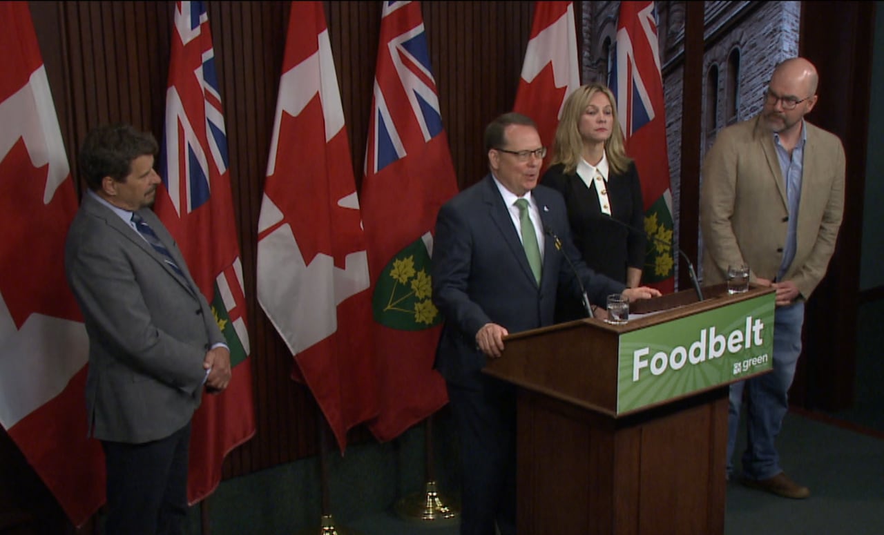 Four people stand on a stage. One man is behind a podium while the others listen.