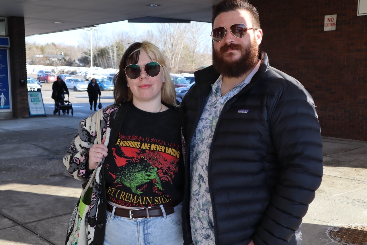 Photo shows a woman and man who are both wearing sunglasses standing beside each other.