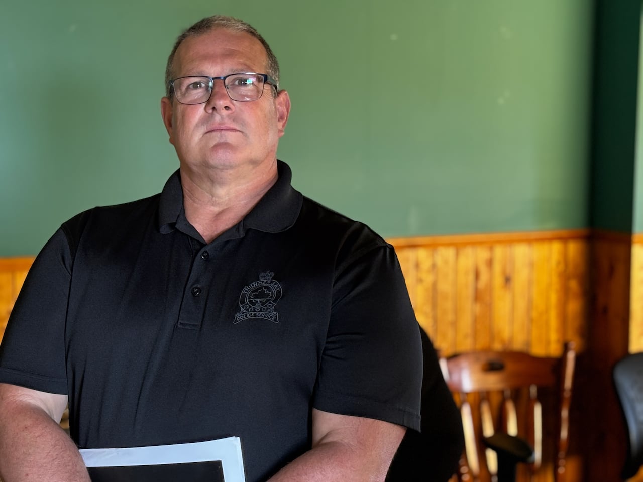 A police officer in glasses and a t-shirt, holding files, looks at the camera with a serious expression on on his face.