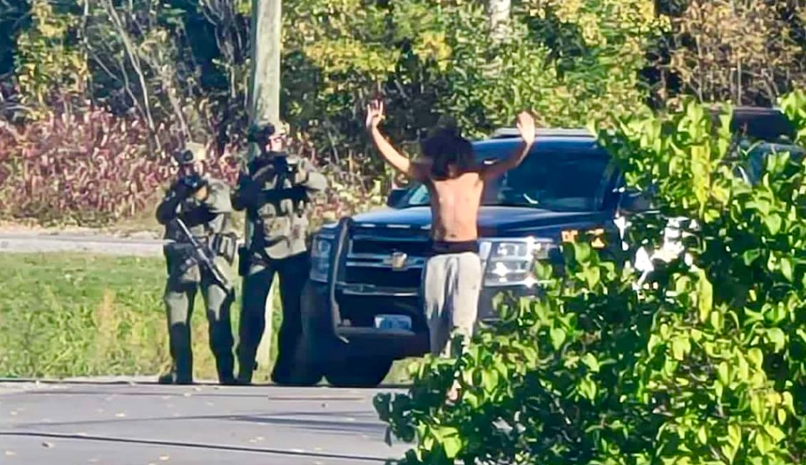 A man with his hands raised surrenders to two police officers behind him, both pointing rifles at him.