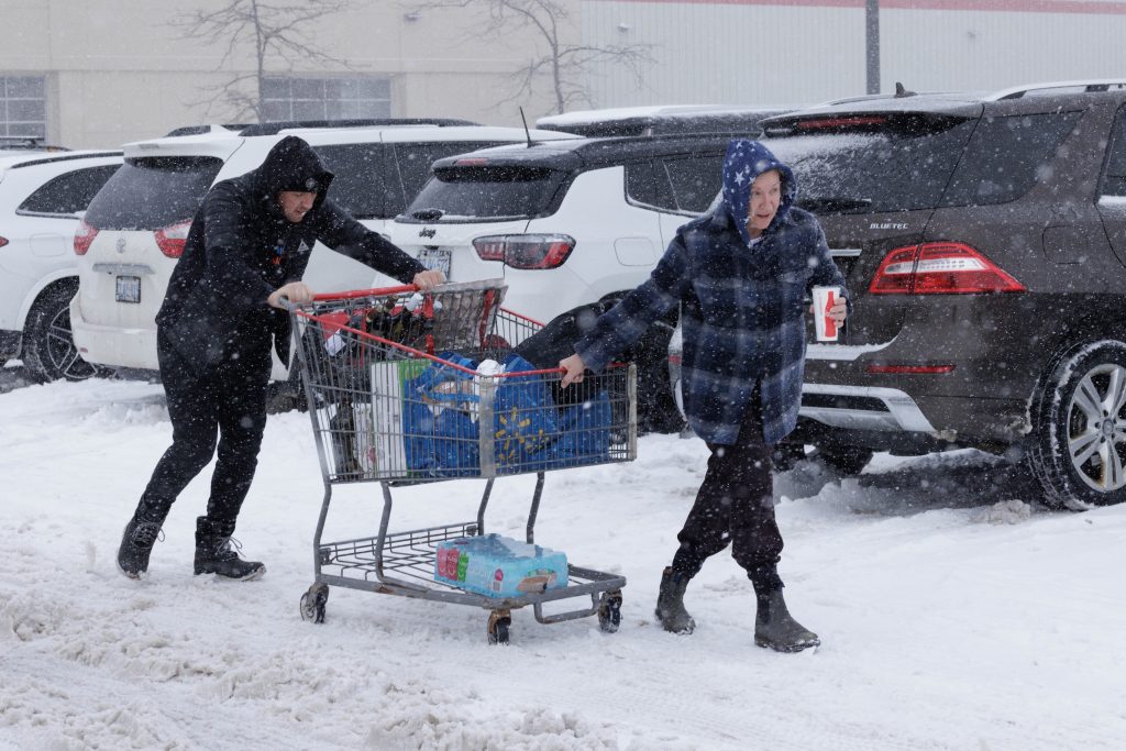 Two people lead a full shopping cart through a snowy parking lot.