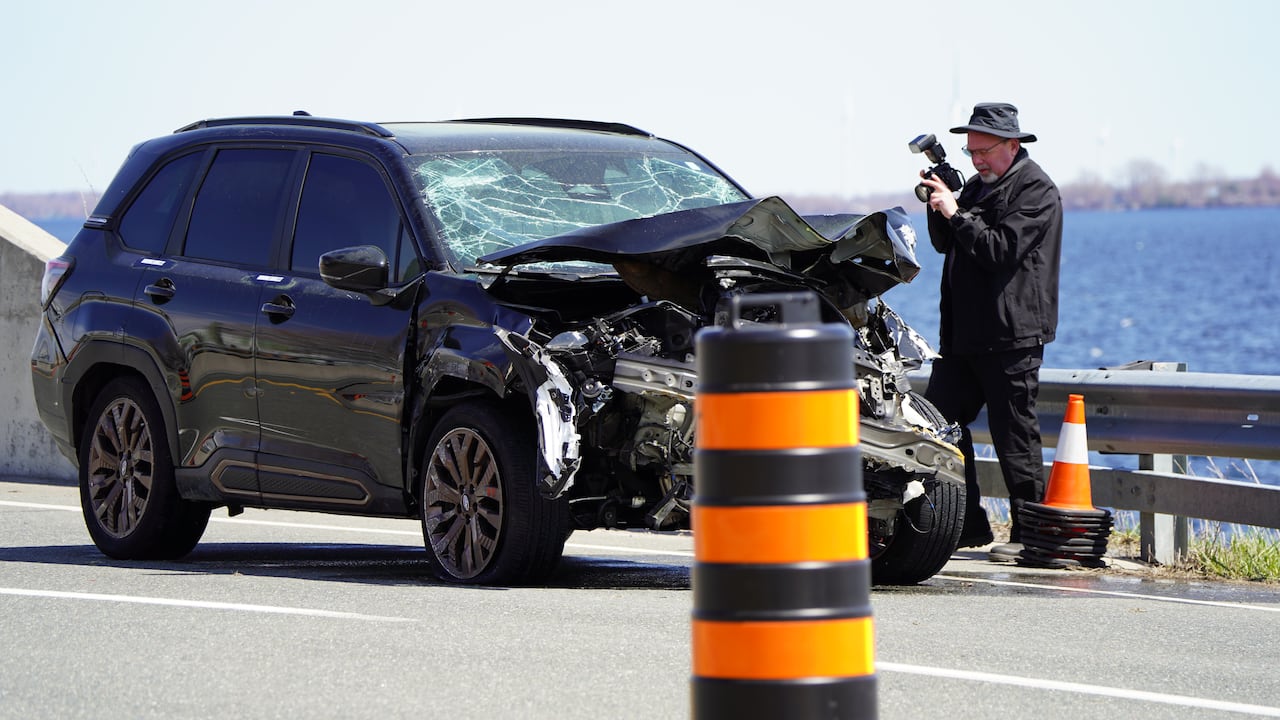 A man in a black Tilley hat and grey shirt uses a camera to take a photo of a black SUV with a smashed windscreen and front end.
