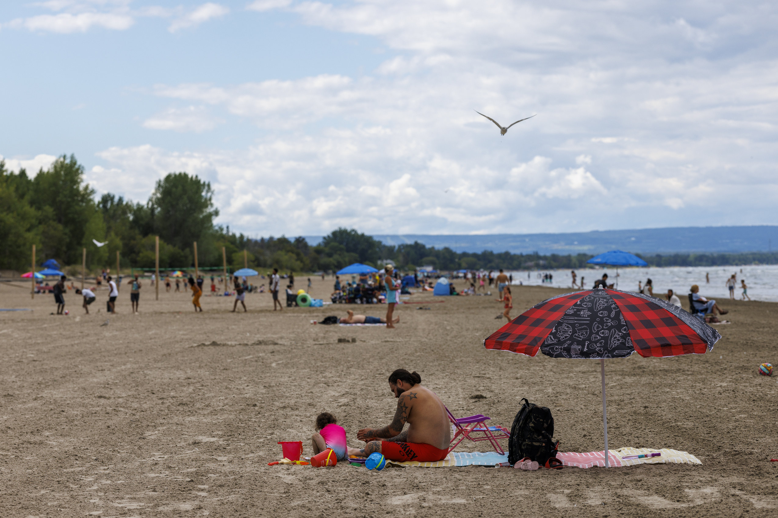 A man and a young girl sit on the sand with a beach umbrella next to them on Wasaga Beach. In the background, other beachgoers enjoy the summer day.