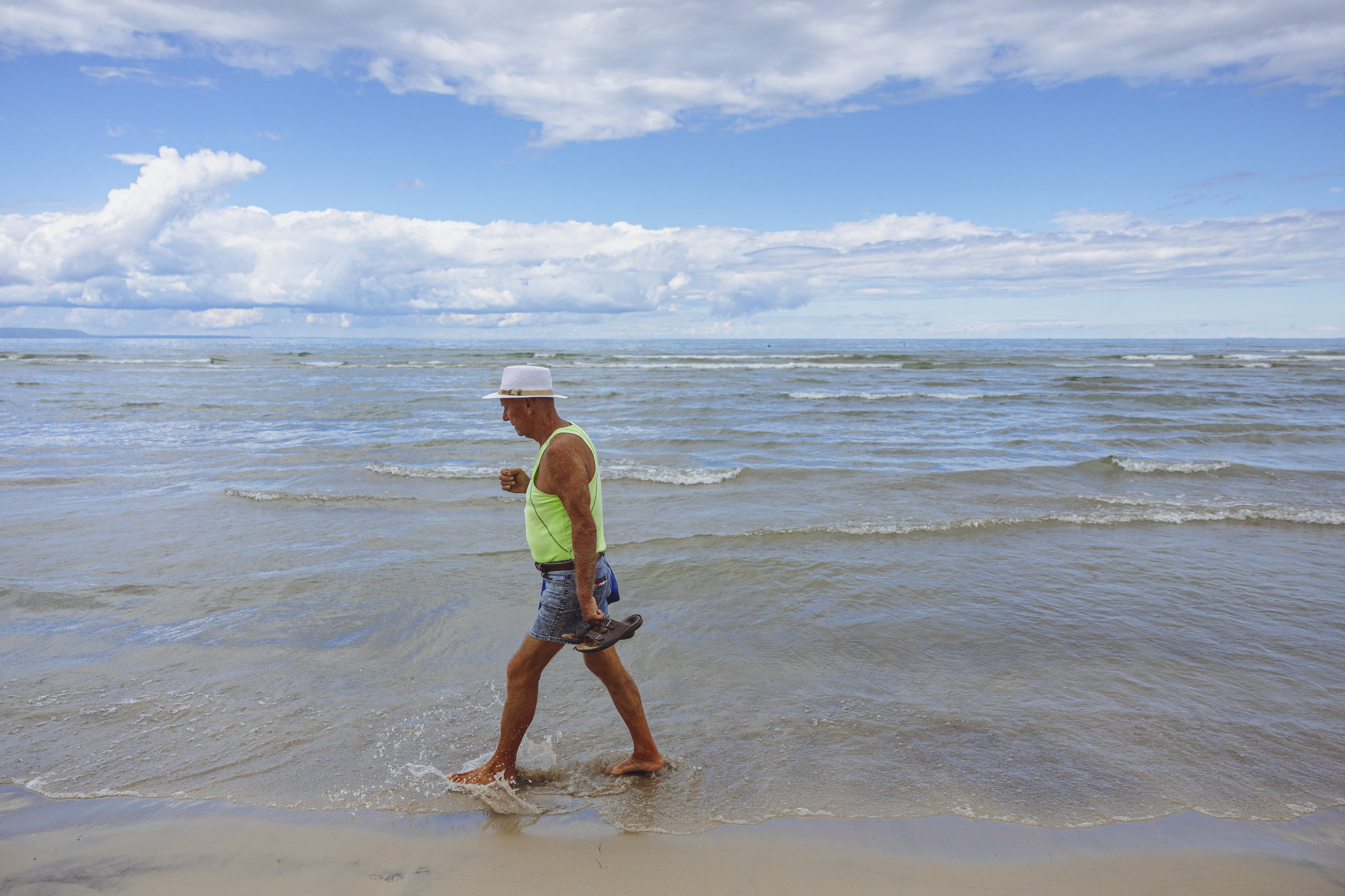 A man walks barefoot along the shore of Wasaga Beach, with the waves reaching his ankles. An expansive lake and sky extend behind him.