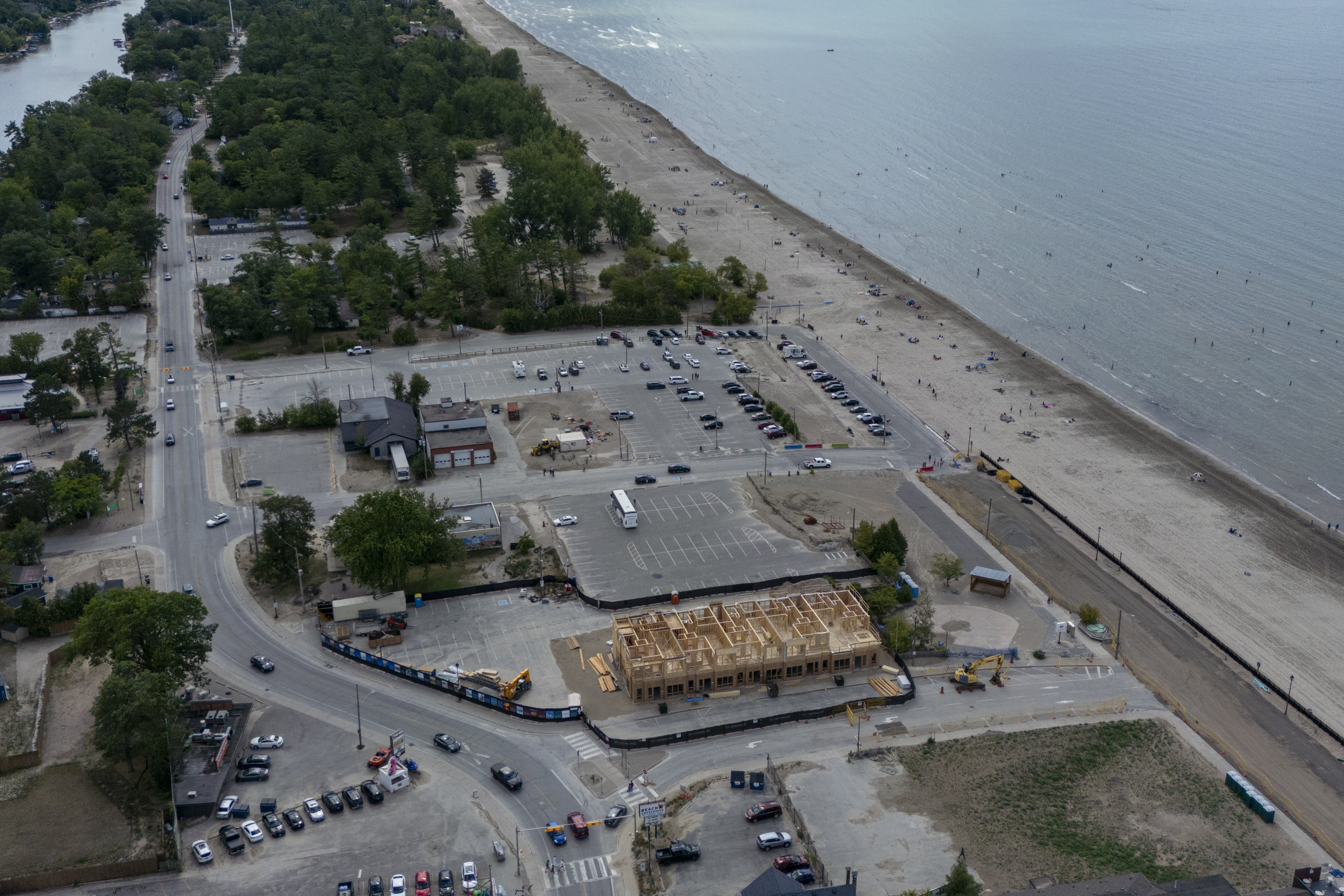 An aerial view of Wasaga Beach. On the right, Lake Huron and the sandy shoreline are dotted wit bathers. On the left, a road, parking lot and wood-framed building under construction.