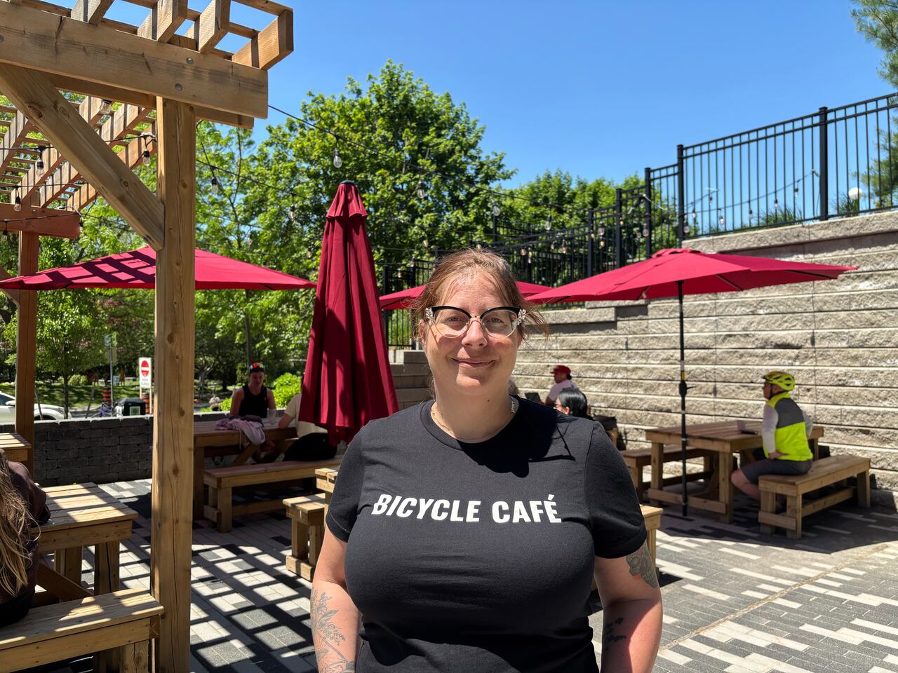 Alicia Berkelmans, executive chef and general manager of the London Bicycle Café, stands on the café's outdoor patio on a sunny day. Wooden picnic tables with red umbrellas and string lights surround the patio, while customers sit in the background.