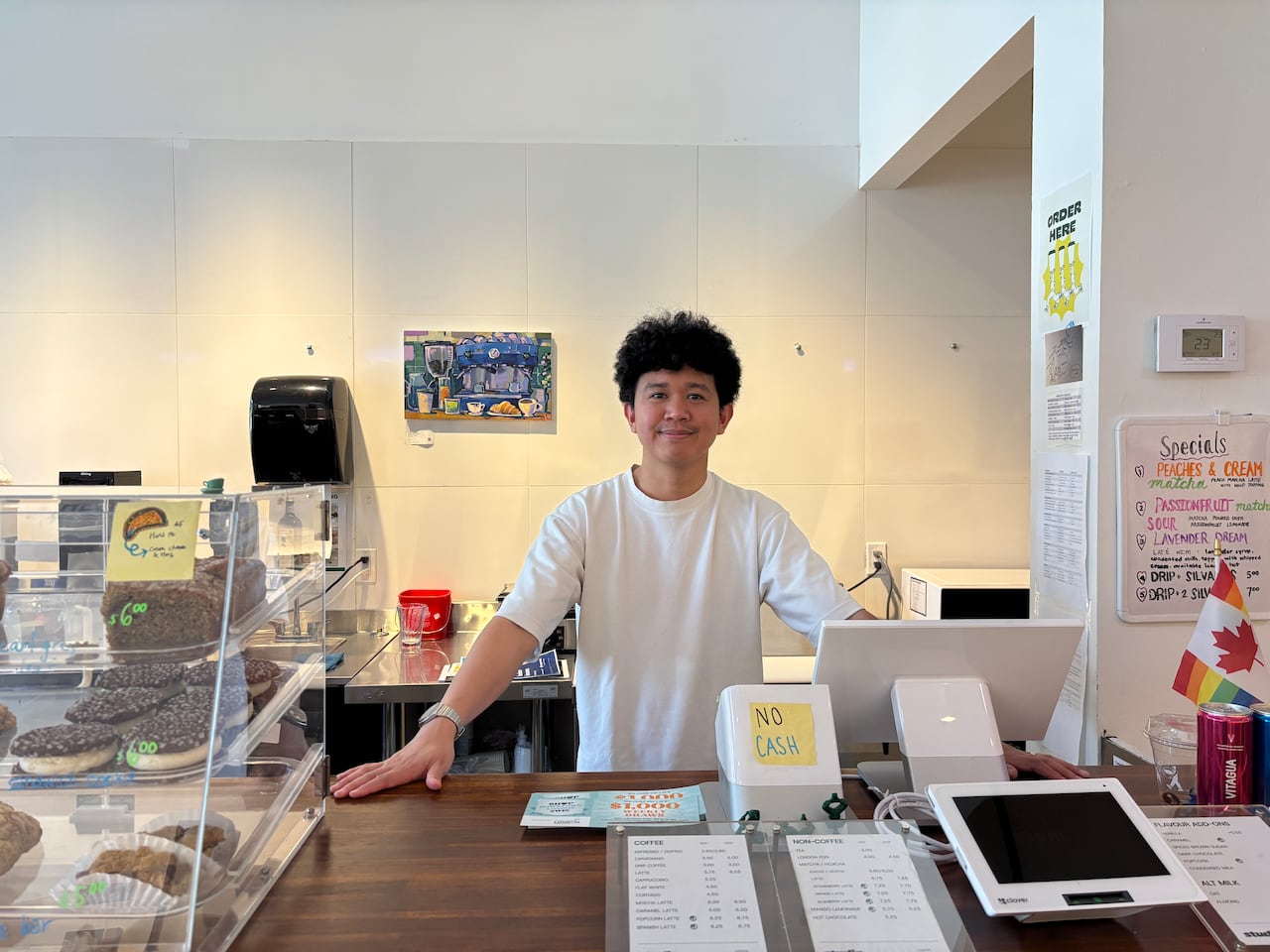 Ashton Samson stands behind the counter at Studio Coffee in downtown London. A glass display case filled with baked goods sits in the foreground, while coffee menus, artwork and café equipment are visible inside the bright, modern coffee shop.