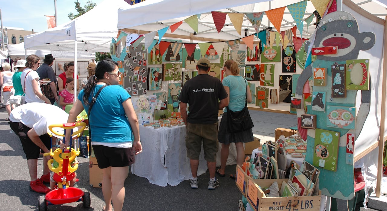 People walk by a colourful booth full of wooden art pieces that depict sock monkeys and other animals
