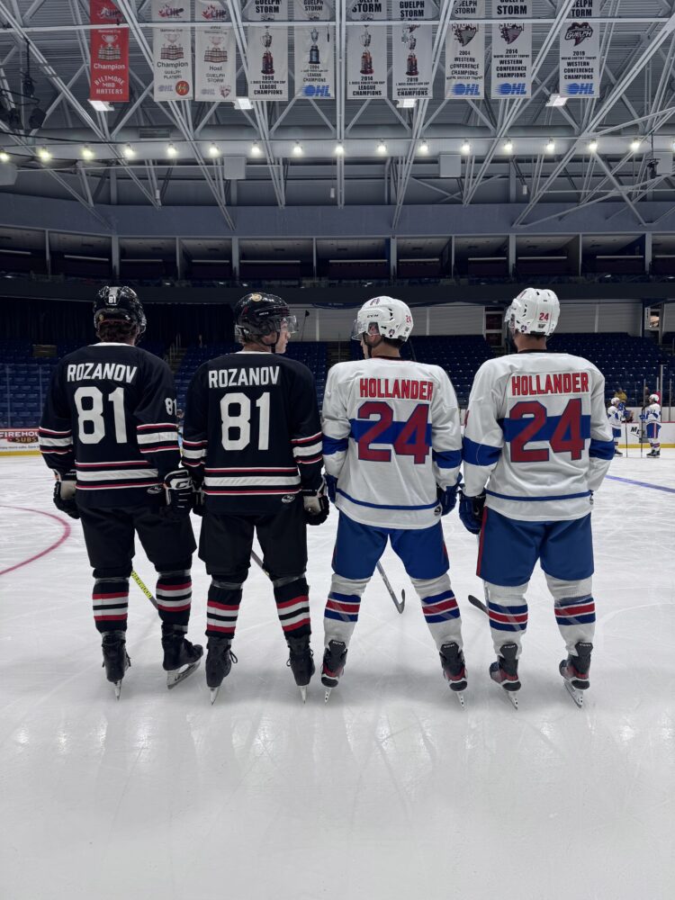 A group of four hockey players wearing black and white uniforms stand with their backs facing cameras showing their names on ice while holding sticks.