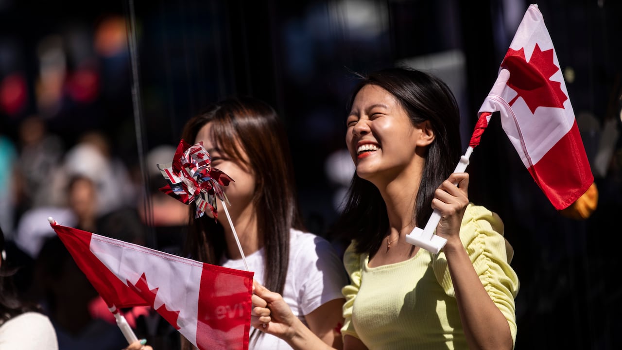 Two women are laughing as they hold Canada flags. One of them is holding a pinwheel with various countries' flags.