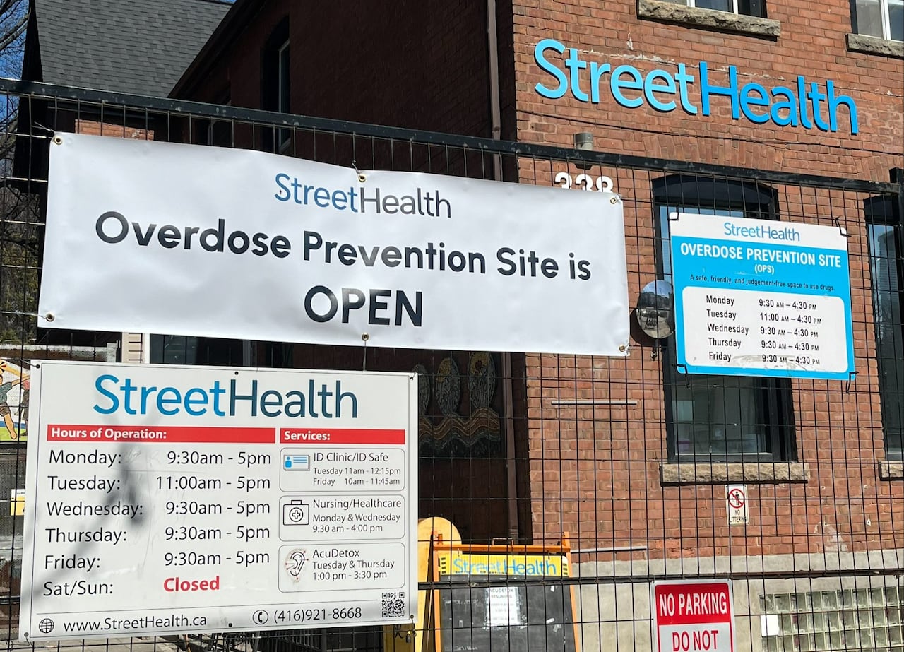 A sign attached to a fence outside a building for StreetHealth reads, "Overdose Prevention Site is OPEN", with their hours below.