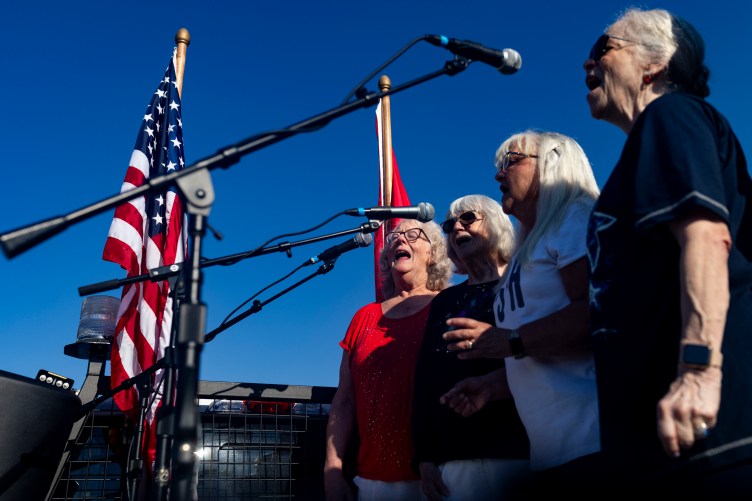 Four women stand side by side singing into microphones outdoors during a daytime event. Flags flank them in background against clear deep blue sky.