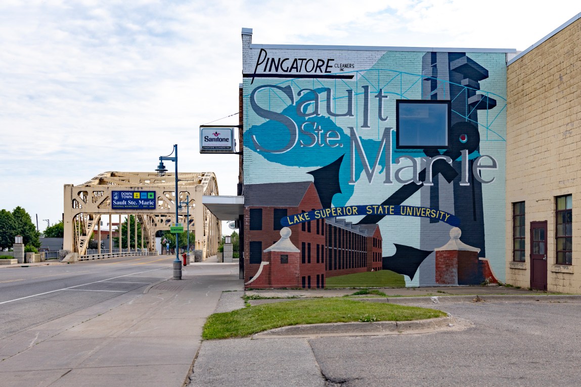 A street-level view shows building adorned large mural includes phrases Pingatore Cleaners Inc Lake Superior State University background light-yellow steel spans road partly cloudy sky.