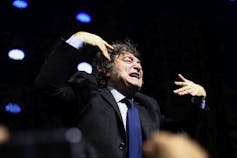 A man with shaggy brown hair grimaces while gesturing with his hands.