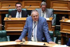 An older man in a blue suit speaks in parliament.