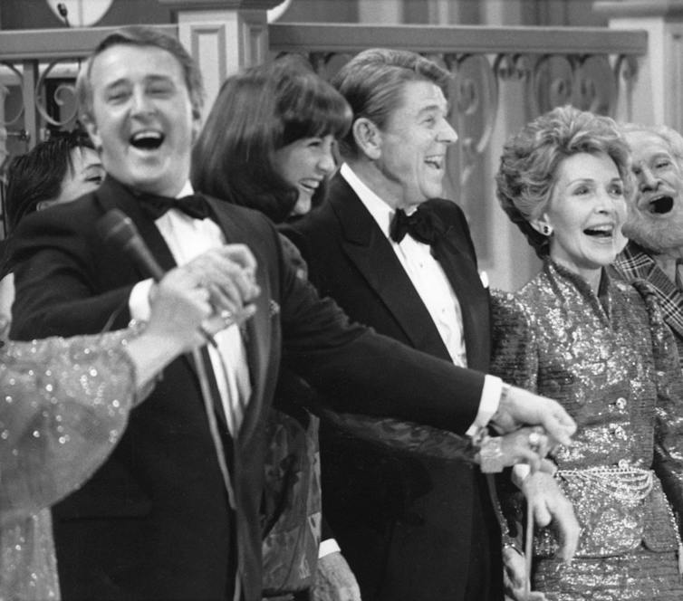 politicians are in tuxedos and their wives in gowns as they grasp hands and sing on stage