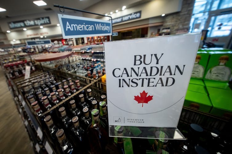 A sign that says 'Buy Canadian Instead' sits beside shelves of whiskey in a liquor store