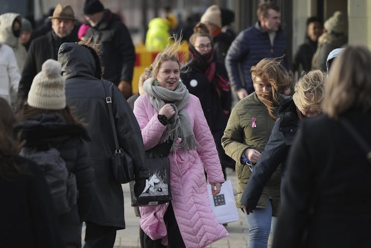 A smiling woman in a pink winter coat smiles as she walks along a city street.
