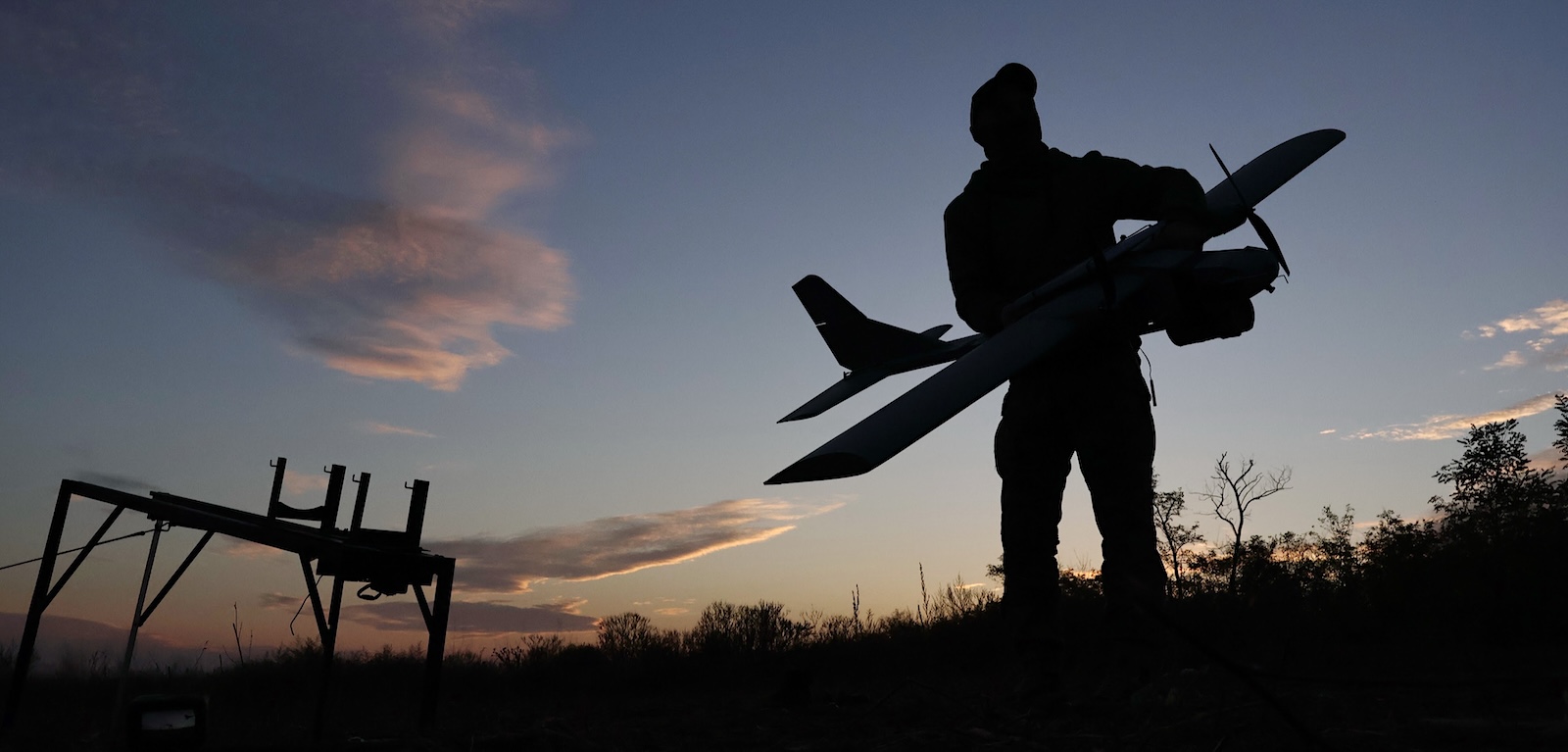 A figure holding a small drone plane is silhouetted against a blue sky with some wispy clouds.